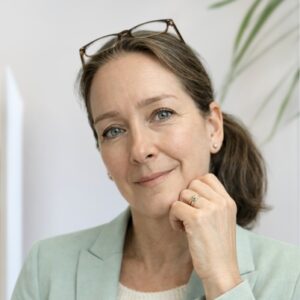 A woman with light brown hair tied back, wearing glasses on her head and a light green blazer, smiles gently while resting her chin on her hand. A plant is visible in the background.