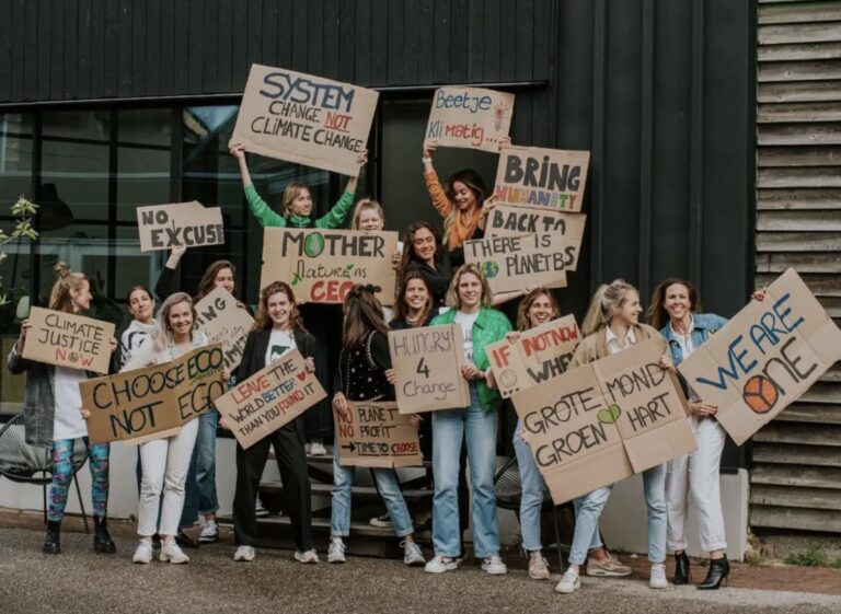 A group of young people stand together outside a building, smiling and holding handmade protest signs about climate change, such as “System change not climate change,” “Mother Nature is CEO,” and “We are one.”.