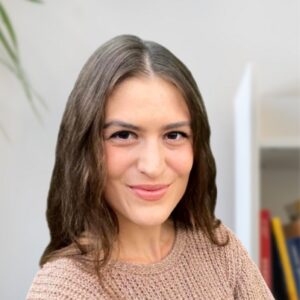 Een vrouw met lang bruin haar en een beige gebreide trui glimlacht naar de camera. De achtergrond is onscherp, met links een plant en rechts planken met boeken.