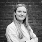 Eine Frau mit langen, hellen Haaren und einem neutralen Gesichtsausdruck steht vor einer Backsteinmauer und trägt ein helles Sweatshirt. Das Bild ist in schwarz-weiß gehalten.