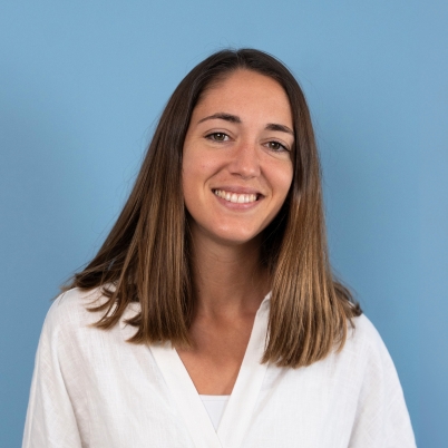 A woman with straight, shoulder-length brown hair is smiling at the camera. She is wearing a white shirt and is posed in front of a plain light blue background.