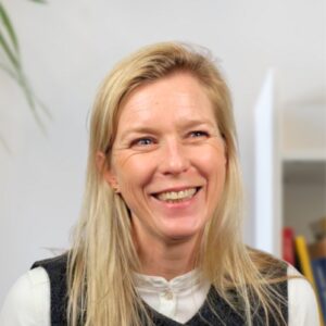 A woman with long blonde hair and a white blouse smiles warmly while looking slightly to the side, sitting indoors against a light background with a few blurred objects and a plant visible.