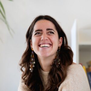 A woman with long brown hair and beaded earrings smiles brightly, looking slightly up. She wears a cream-colored sweater, and the background is softly blurred with hints of green leaves and a door.