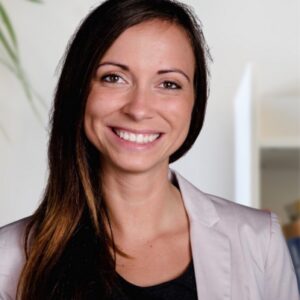 Eine Frau mit langen braunen Haaren, die einen hellen Blazer und ein schwarzes Oberteil trägt, lächelt in einer hellen Innenumgebung in die Kamera.