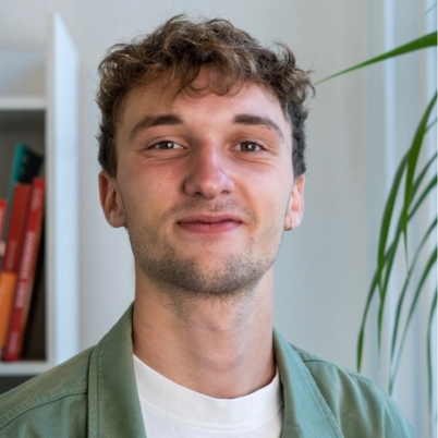 A young man with curly light brown hair and a light beard smiles at the camera. He is wearing a green jacket over a white shirt. There is a plant and a white bookshelf with books in the background.