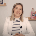 Eine Frau mit blondem Haar, die einen weißen Blazer und ein gestreiftes Oberteil trägt, sitzt an einem Tisch und hat ein Mikrofon am Revers befestigt. Im Hintergrund sind Regale mit Dekorationsartikeln zu sehen.