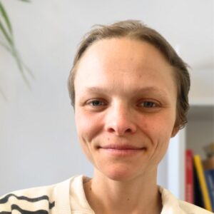 A person with short light brown hair and light skin smiles gently at the camera. They wear a white top with dark stripes. The background is softly blurred, showing part of a bookshelf and a green plant.