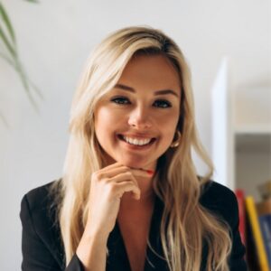 A smiling woman with long blonde hair and a black top sits indoors, resting her chin on her hand. The background is softly blurred, with a hint of plants and bookshelves visible.