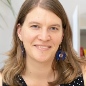 A woman with straight, light brown hair and blue earrings smiles warmly at the camera. She is wearing a black top with white polka dots and has a lip piercing. The background is softly blurred.
