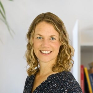 A woman with shoulder-length wavy blonde hair smiles at the camera. She is wearing a dark patterned blouse and is standing indoors with a blurred background, including books and a plant.