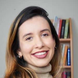 A woman with long dark hair and highlights, wearing a beige turtleneck sweater, smiles in front of a blurred bookshelf.