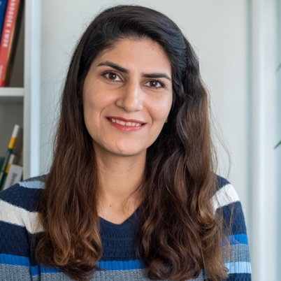 A woman with long brown hair and a striped blue sweater smiles at the camera, sitting indoors near a white bookshelf.