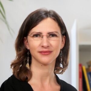 A woman with shoulder-length brown hair, wearing glasses and dangling earrings, smiles softly. She is dressed in a black top, with a blurred bookshelf and white wall in the background.