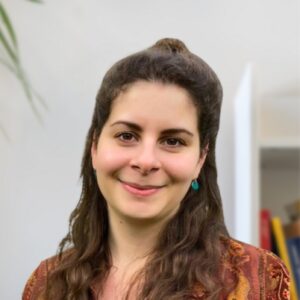 Eine Frau mit langen braunen Haaren und türkisfarbenen Ohrringen lächelt sanft in die Kamera. Sie trägt ein gemustertes Oberteil, und im hellen Hintergrund sind ein verschwommenes Bücherregal und eine Pflanze zu sehen.