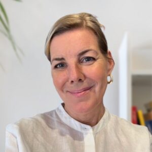 A woman with short blonde hair and gold hoop earrings, wearing a white blouse, smiles at the camera in a bright room with a plant and bookshelf in the background.