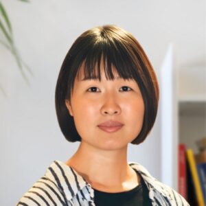 A woman with straight, chin-length dark hair and bangs, wearing a black top and a light-colored, striped shirt, stands indoors with a neutral expression. Blurred books and a plant are visible in the background.