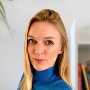 Eine Frau mit langen blonden Haaren und blauen Augen, die einen blauen Rollkragenpullover trägt, blickt in die Kamera, vor einem unscharfen Innenhintergrund mit einer weißen Wand und einigen Büchern.