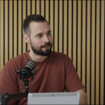A man with a beard wearing a rust-colored shirt sits in front of a microphone and tablet, speaking about Leadership in the Era of AI, with a wooden slat wall in the background.