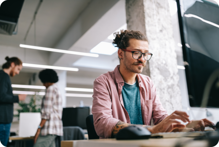 Een man met bril en een knotje typt op een toetsenbord aan een bureau in een modern kantoor. Twee mensen praten op de achtergrond. De werkruimte is helder met natuurlijk en kunstmatig licht.