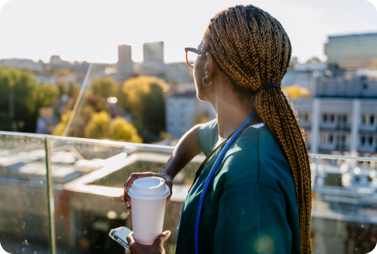 Eine Person in medizinischem Kittel mit geflochtenem Haar hält eine Kaffeetasse und ein Telefon in der Hand und blickt an einem sonnigen Tag von einer Dachterrasse auf eine Stadtlandschaft.