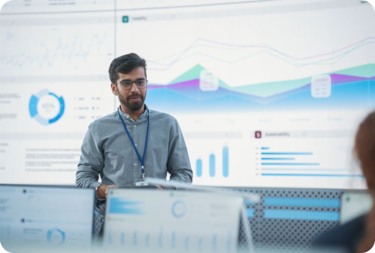 Een man met een bril en een badge presenteert gegevens in een modern kantoor, staande voor grote digitale schermen met grafieken, diagrammen en datavisualisaties.