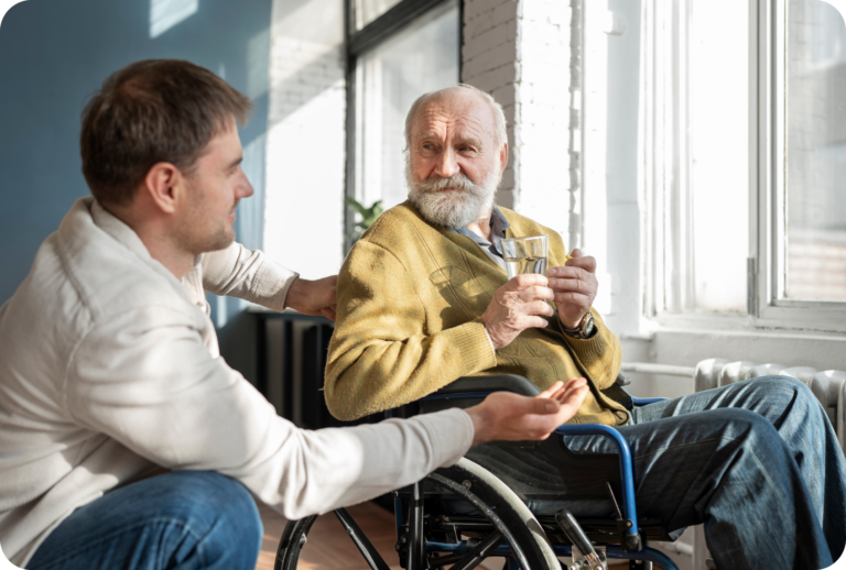 Ein junger Mann unterhält sich mit einem älteren bärtigen Mann im Rollstuhl, der mit einem Glas Wasser in der Hand an einem sonnigen Fenster in einem hellen Raum sitzt.