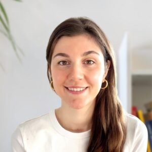 A young woman with long brown hair, wearing gold hoop earrings and a white shirt, smiles at the camera. The background is softly lit with blurred indoor details and a hint of a green plant.
