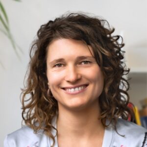 Eine Frau mit lockigem braunem Haar und einem freundlichen Lächeln sitzt in einem Haus und trägt ein helles Oberteil. Der Hintergrund ist leicht unscharf und zeigt einen Teil einer Pflanze und Regale.