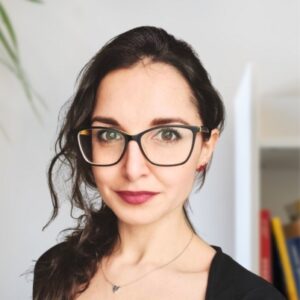 Eine Frau mit langen, dunklen, gewellten Haaren, die eine schwarze Brille und ein schwarzes Oberteil trägt, steht in einem Innenraum in weichem Licht. Im Hintergrund sind eine Tür und verschwommene Gegenstände zu sehen. Sie blickt direkt in die Kamera und lächelt leicht.