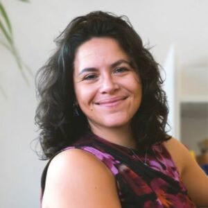 Eine Frau mit gewelltem dunklem Haar und einem ärmellosen geblümten Oberteil lächelt warm, während sie in einem Haus sitzt.