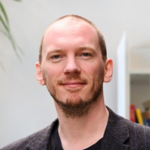 A man with a light complexion and short, light brown hair and beard, wearing a dark blazer over a black shirt, stands indoors and smiles gently at the camera. The background is softly lit and out of focus.