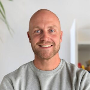 Ein lächelnder Mann mit Glatze und gestutztem Bart, der ein hellgraues Sweatshirt trägt, steht in einem hellen Raum mit einer weißen Wand und einer teilweise sichtbaren Pflanze im Hintergrund.