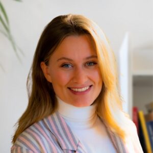 A woman with blonde hair and blue eyes, wearing a white turtleneck and a light-colored striped blazer, smiles warmly at the camera. The background is softly lit with some books and a plant visible.