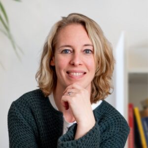 Eine Frau mit blondem, schulterlangem Haar und blauen Augen lächelt in die Kamera und stützt ihr Kinn auf ihre Hand. Sie trägt einen strukturierten dunkelgrünen Pullover und ein weißes Oberteil. Der Hintergrund ist sanft verschwommen.