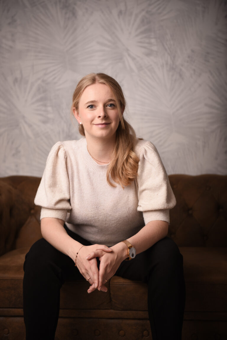 Eine Frau mit blondem Haar, die ein helles Oberteil mit Puffärmeln und eine schwarze Hose trägt, sitzt leicht lächelnd auf einem braunen Sofa vor einem hell gemusterten Hintergrund.