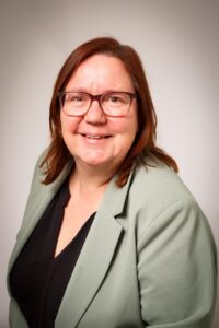 Een vrouw met schouderlang bruin haar en een bril lacht naar de camera. Ze draagt een lichtgroene blazer over een zwart topje, met een effen lichte achtergrond.