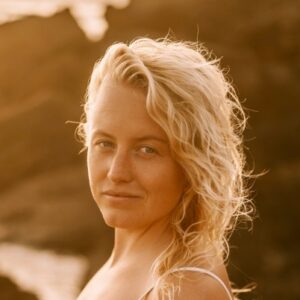 Een vrouw met golvend blond haar staat buiten in warm, gouden zonlicht en kijkt met een lichte glimlach naar de camera. De achtergrond is wazig, met donkere rotsen en een vleugje water.