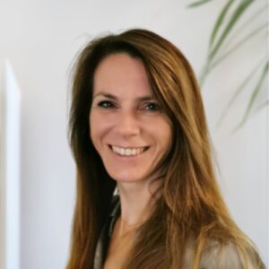 Een vrouw met lang bruin haar glimlacht warm naar de camera tegen een helderwitte achtergrond met een vleugje groen gebladerte.