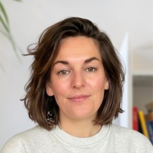 Een vrouw met schouderlang bruin haar en een lichtgrijze trui kijkt naar de camera en glimlacht lichtjes. De achtergrond is zachtjes wazig met wat planten en planken zichtbaar.