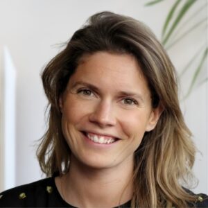 Een vrouw met schouderlang lichtbruin haar lacht naar de camera. Ze draagt een zwart topje en poseert binnenshuis, met een onscherpe achtergrond met een groene plant.