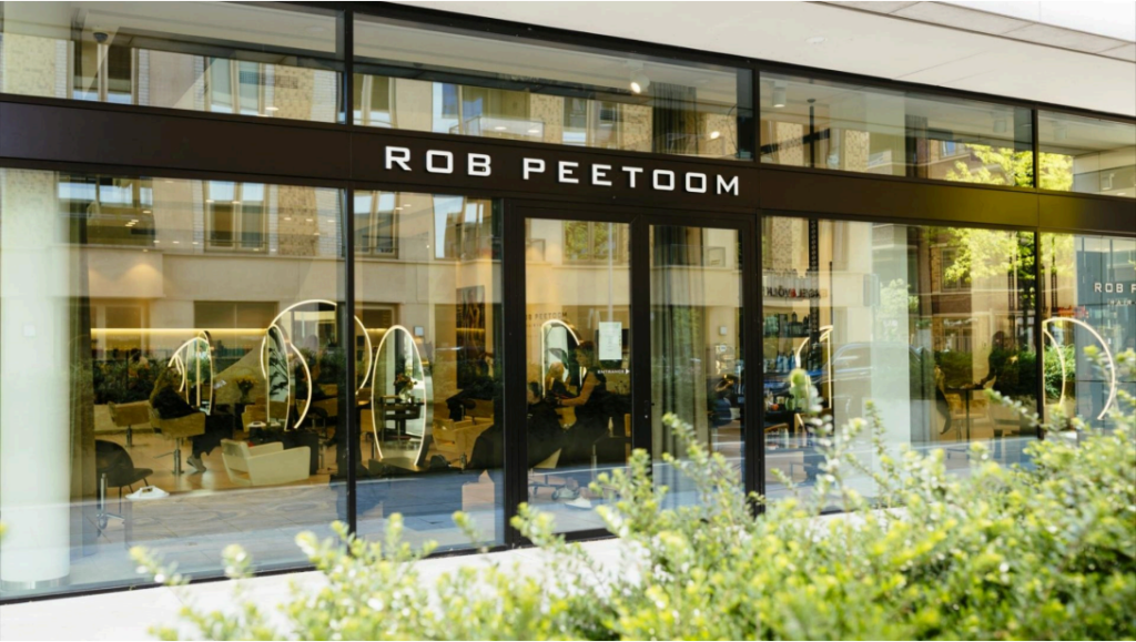 The image shows the exterior of a modern hair salon with large glass windows and the name ROB PEETOOM displayed above the entrance. Inside, several salon chairs and mirrors are visible. Greenery is in the foreground.