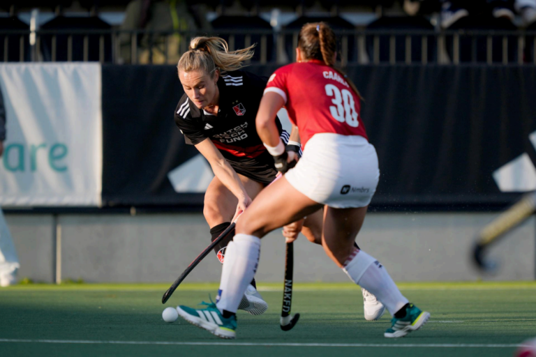 Two female field hockey players compete for the ball on a green field. The player in black is dribbling, while the player in red, number 38, is defending. Spectators are visible in the blurred background.