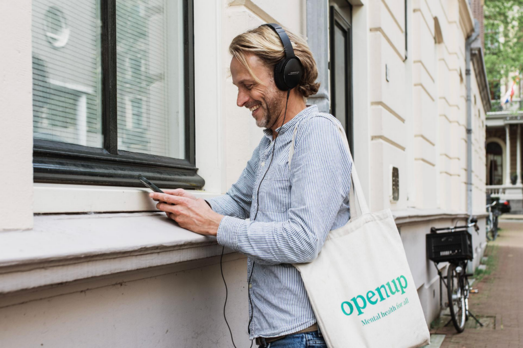 A smiling man with headphones stands outside by a building, using his smartphone. He carries a white tote bag with the text “openup Mental Health for all.” A bicycle is parked behind him on the sidewalk.