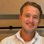 Een man met kort, lichtbruin haar glimlacht terwijl hij zit. Hij draagt een wit overhemd en poseert tegen een beige en bruine achtergrond.