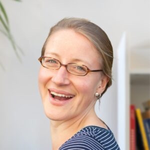 Een vrouw met lichtbruin haar, een bril en een gestreept shirt lacht naar de camera. Er zijn wat boeken en een plant zichtbaar in de zacht vervaagde achtergrond.