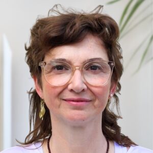 Een vrouw met golvend bruin haar, een bril met helder montuur en gouden oorbellen, glimlacht zachtjes naar de camera. Ze is gekleed in een licht topje en verschijnt binnenshuis met een groene plant op de achtergrond.