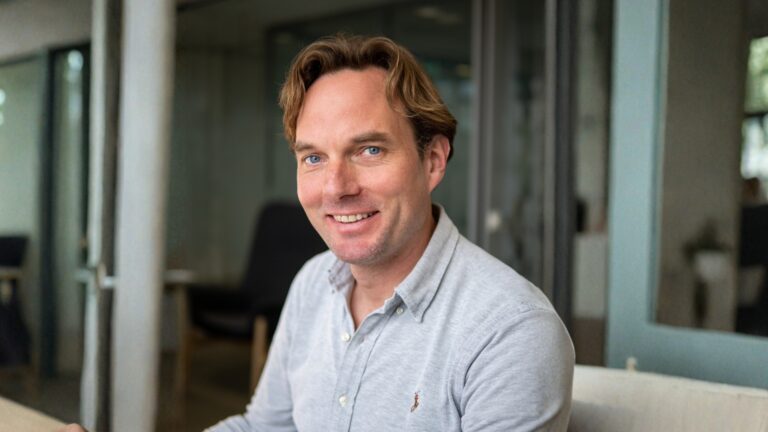 A man with light brown hair and blue eyes, wearing a light gray button-up shirt, sits indoors smiling at the camera. The background shows a modern office setting with glass walls and furniture.