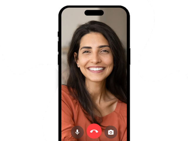 A woman with long dark hair and a warm smile appears on a smartphone screen during a video call, wearing an orange top. Video call icons are visible at the bottom of the screen.