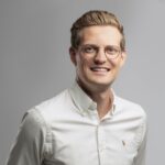 Ein lächelnder Mann mit kurzen blonden Haaren und Brille, der ein weißes Hemd mit Knöpfen und einem kleinen gestickten Logo trägt, posiert vor einem schlichten grauen Hintergrund.