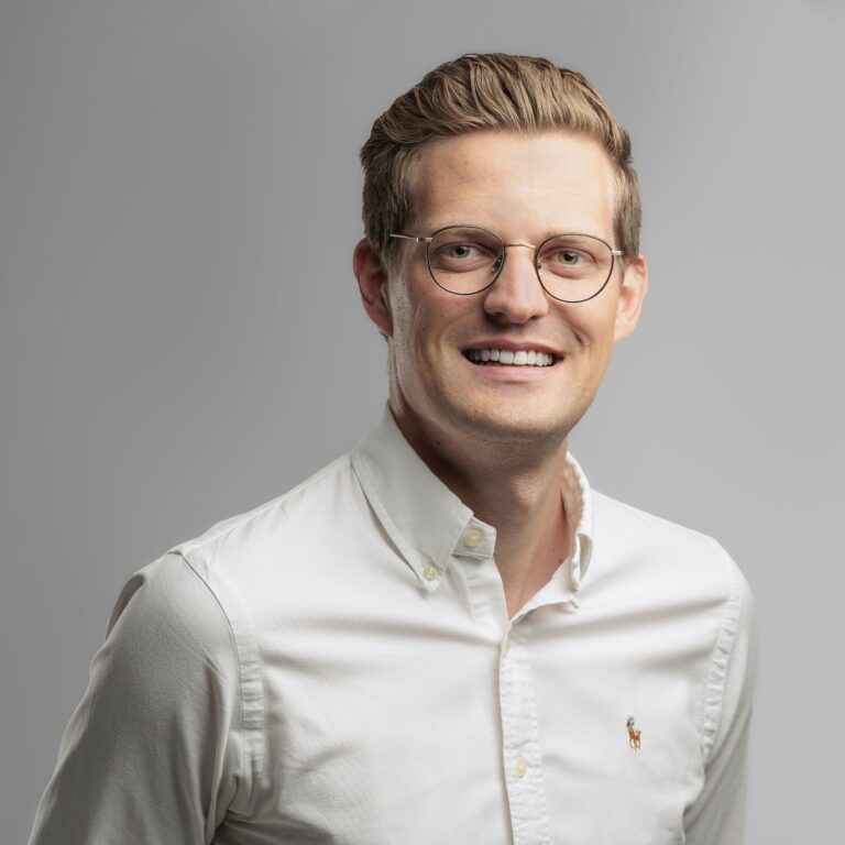 Ein lächelnder Mann mit kurzen blonden Haaren und Brille, der ein weißes Hemd mit Knöpfen und einem kleinen gestickten Logo trägt, posiert vor einem schlichten grauen Hintergrund.